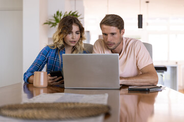 Couple sitting at home kitchen table using laptop on woven placemat with smartphone tablet and mug
