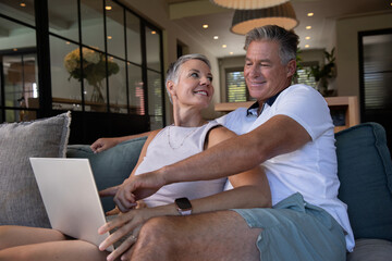 Senior couple sitting on teal couch under pendant lamp in home living room, using laptop