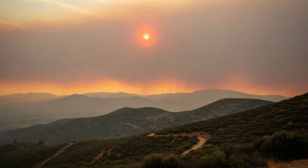 Fototapeta premium A hazy landscape with a winding dirt road on a hillside, shrouded in smoky haze under a reddish-orange sunset, indicating pre-disaster conditions and environmental risk. Perfect for public