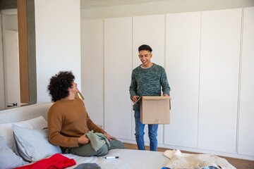Male roommates unpacking and organizing clothes in bright bedroom using cardboard box and marker