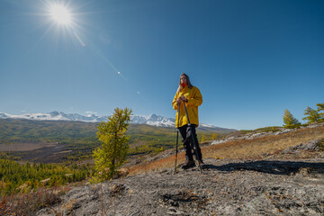 Female traveler in a yellow jacket stands on a rocky mountain ridge with trekking poles, admiring the panoramic view of snow-capped peaks and an autumn forest valley under a bright sun. © ANDREY