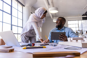 Diverse coworkers reviewing architectural plans on desk in open office with tablet and scale model