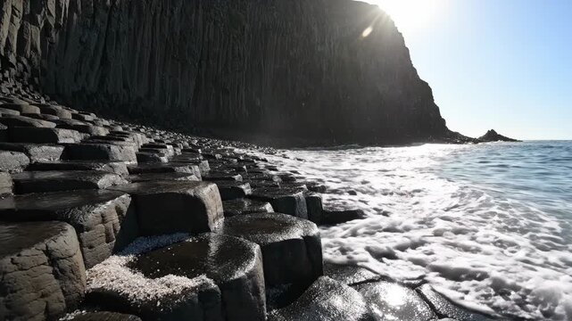Sunlight streams over hexagonal rock formations on a dramatic coast Waves crash against the dark basalt columns creating a mesmerizing natural spectacle