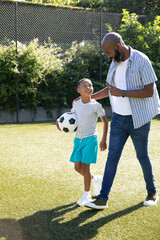 African American father and son walking on grass pitch near fence pointing and carrying soccer ball