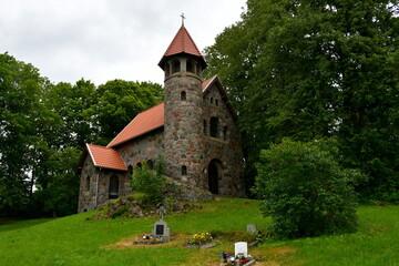 Obraz premium A view of an unique old church made entirely out of stones, boulders, and rocks with a red tile roofing standing in the middle of a meadow of pastureland, seen on a sunny summer day in Poland