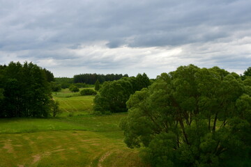 A view of a vast meadow, lawn or pastureland surrounded with trees, shrubs, flowers and other flora seen next to a biking path, some wooden benches and bike stands in summer in Poland