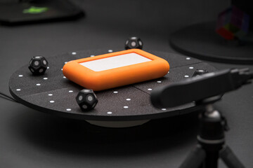A close-up of an orange gadget resting on a black circular platform surrounded by small dice, suggesting a tech or gaming scene in a studio setting.