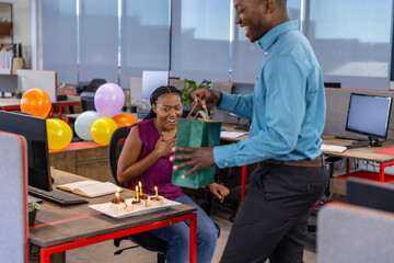 African american coworkers celebrating birthday at office desk with cake balloons, gift bag