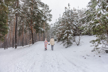 Young woman and child walking hand in hand along snowy forest path during winter snowfall, peaceful outdoor family moment with pine trees, fresh snow, cold weather atmosphere 