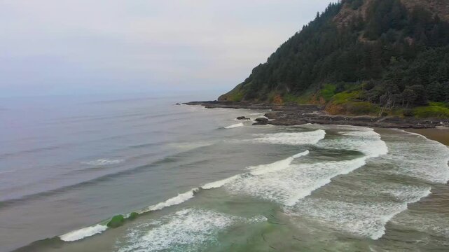 An aerial, wide shot view of the dramatic and rugged coastline at Cape Perpetua, Oregon. Large waves crash onto the expansive, rocky beach and a steep, forested bluff rises from the shore under an ove