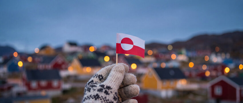 Hand in a knitted wool mitten holding a small Greenland flag against a backdrop of blurred colorful houses and city lights at twilight in the Arctic.
