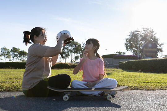 Asian mother removes a safety helmet from her young daughter while sitting outdoors after activity, showing parental care, child safety, and family bonding.