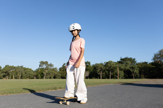 Asian girl skateboarding alone outdoors while wearing a safety helmet, showing child safety, outdoor activity, and active childhood lifestyle.