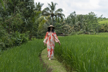Woman walking along the green rice fields wearing long red and white dress and Asian conical hat in Bali Indonesia