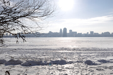 Snow-covered riverbank in the foreground with a large city skyline across a wide frozen river. Bright winter day with clear sky, icy river surface and urban winter landscape