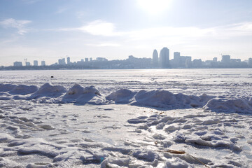 Snow-covered riverbank in the foreground with a large city skyline across a wide frozen river. Bright winter day with clear sky, icy river surface and urban winter landscape