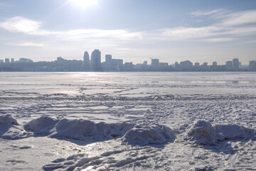 Snow-covered riverbank in the foreground with a large city skyline across a wide frozen river. Bright winter day with clear sky, icy river surface and urban winter landscape