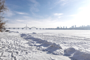 Snow-covered riverbank in the foreground with a large city skyline across a wide frozen river. Bright winter day with clear sky, icy river surface and urban winter landscape