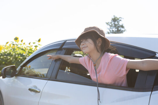 Asian girl with arms outstretched leaning near a car window during a family trip, showing childhood joy, curiosity, and relaxed travel lifestyle.