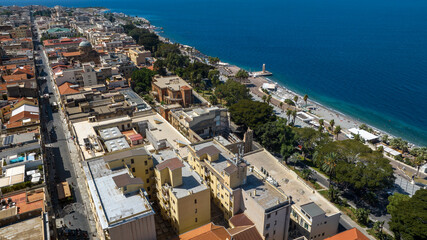 Aerial view of a coastal city featuring historic buildings with red roofs, a long tree-lined promenade, and a turquoise bay next to a lush green urban park. It's the Reggio Calabria waterfront, Italy.