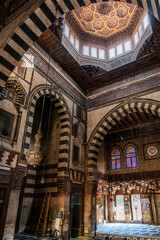 Interior view of Sultan Al-Ashraf Qaytbay Mosque and mausoleum. Features intricate arches, striped walls, and a decorative dome. Soft light filters through stained glass windows.
