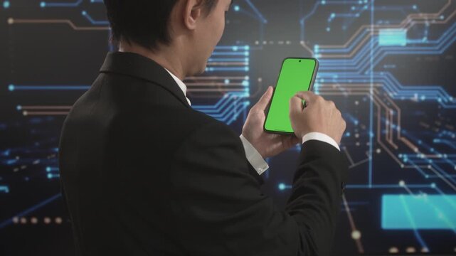 Rear view of Asian businessman using smartphone with green screen display against futuristic digital circuit background, representing mobile application testing, technology innovation
