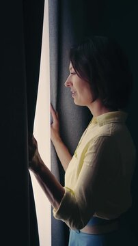 Woman opens curtains in a living room to let in morning light and fresh air.