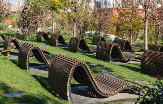 Modern park with curved wooden benches on green grass, surrounded by trees and city buildings in background. The recreation area is on mound in Oblaka Park. Krasnodar Public Park or Galitsky Park.