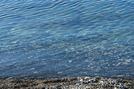 Shallow shoreline with clear blue water gently washing over smooth pebbles and small stones. Ripples and reflections create serene, tranquil scene. Coastline on beach in Gelendzhik