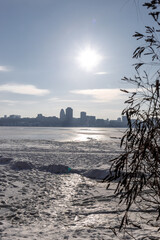 Snow-covered riverbank in the foreground with a large city skyline across a wide frozen river. Bright winter day with clear sky, icy river surface and urban winter landscape