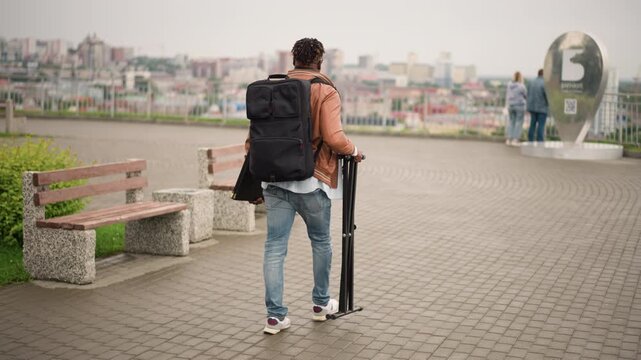 street musician walking through city plaza carrying backpack and folded stand, leather jacket and ripped jeans, empty benches and skyline backdrop, overcast light and contemplative mood