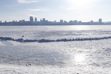 Snow-covered riverbank in the foreground with a large city skyline across a wide frozen river. Bright winter day with clear sky, icy river surface and urban winter landscape