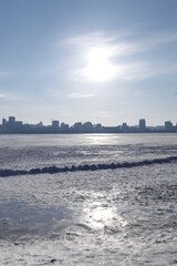 Snow-covered riverbank in the foreground with a large city skyline across a wide frozen river. Bright winter day with clear sky, icy river surface and urban winter landscape