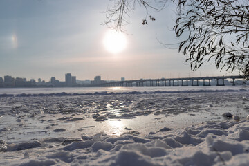 Snow-covered riverbank in the foreground with a large city skyline across a wide frozen river. Bright winter day with clear sky, icy river surface and urban winter landscape