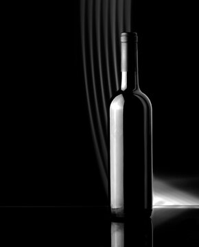 An unopened bottle of wine stands on a glass table against a black background. Black and white image.