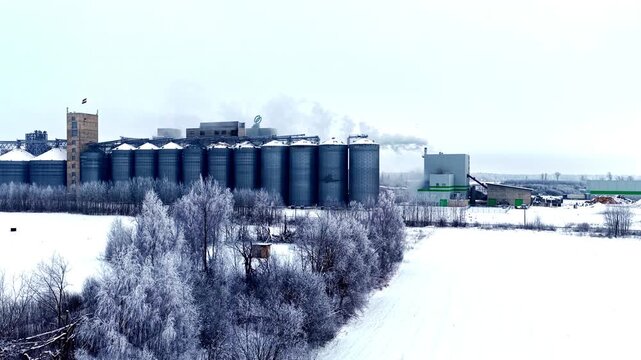 Drone shot rising above large scale grain processing plant with cylindrical metal silos and steam billows in snowy winter landscape near Jelgava town