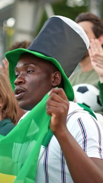 African American young male football fan wearing green hat holding Brazil flag cheering in stadium. Supporter in striped shirt watching match with excitement. Outdoors. Vertical format.