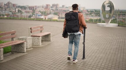 backpacker dj carrying gear along promenade passing benches and railing, panoramic urban skyline, steady stride toward setup location, casual sneakers and focused arrival, creator energy and gig