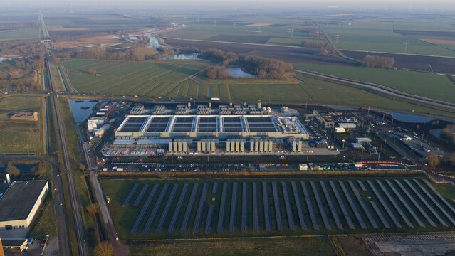 Aerial view of the Google datacenter complex near a canal, contrasting with the adjacent solar panel array under a pale sky, Winschoten, Groningen, Netherlands.