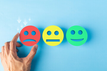 Hand choosing a red sad face paper cut out for negative feedback and bad rating. Concept of customer experience, low satisfaction, service evaluation, and emotional disappointment on blue background.