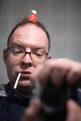 Portrait of a man with glasses taking a photo of himself with a camera, smoking tobacco, and wearing a funny and miniature Santa hat