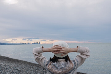 Person standing on a pebble beach facing the ocean with hands behind head, wearing a knitted...