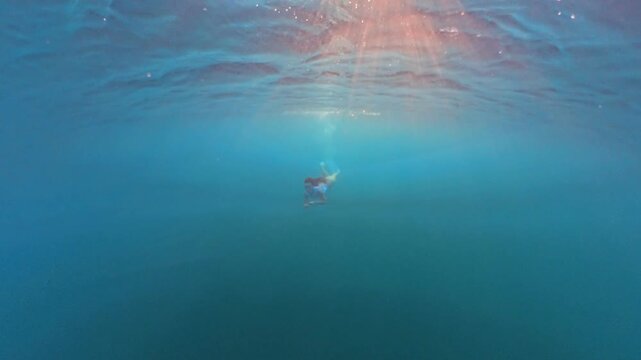 Swimming, freediving, underwater woman gliding through blue ocean water toward bright surface sun rays.
