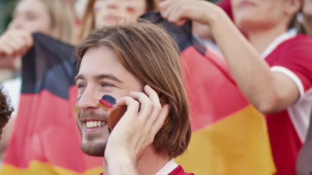 Caucasian male football fan with German flag face paint talking on smartphone in stadium. Man in red shirt making call surrounded by cheering crowd waving German flags at sport game. Outdoors.