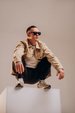 Full body studio shot of a young male model squatting on a white cube. Wearing beige cotton shirt, white t-shirt, black cargo pants and black sunglasses, isolated neutral background.