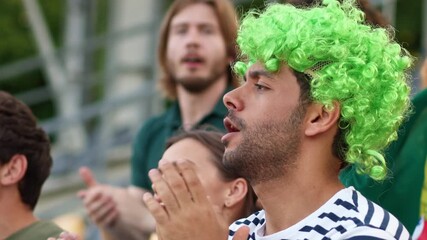 African American handsome male fan wearing curly green wig shouting during football match in stadium. Excited man cheering, supporting national team with crowd. Outdoors. Striped shirt visible.