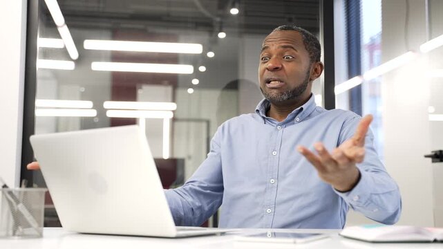 Angry dissatisfied african american businessman arguing talking on video call using laptop sitting at workplace in business office. Frustrated manager emotionally talks about problems expressing anger