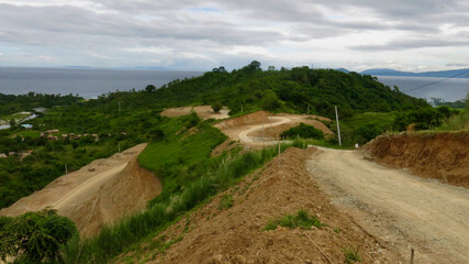 Winding dirt road cut through a green coastal hillside. Mountain landscape shows an unpaved road under construction with tropical vegetation and the sea visible on the horizon.