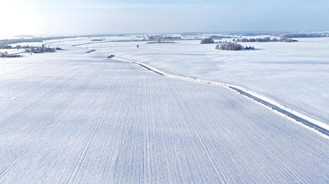 Drone view rising over winter landscape with road curving through flat snow covered farm fields and frozen furrows under bright sunrise sky in rural Latvia