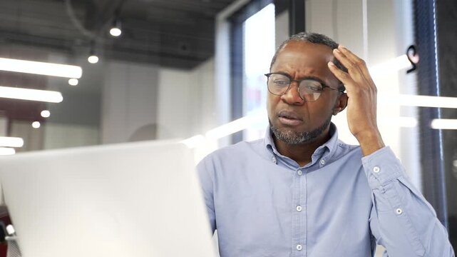Exhausted businessman is bored sitting at workplace in business office. Black man worker is tired from working with laptop computer. Tired overworked employee procrastinates, gets distracted. Close up